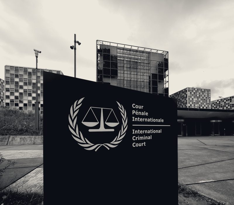 The entrance sign of the International Criminal Court in The Hague, bearing the Court's scales-and-laurel emblem, with its headquarters behind.